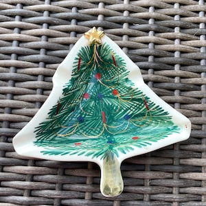 May include: A ceramic Christmas tree-shaped dish with a gold star on top. The tree is painted green with red and blue ornaments, gold garland, and a gold trunk. The dish is on a brown woven surface.