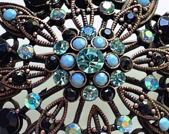 Può includere: Una spilla in metallo color bronzo con un design floreale. La spilla è decorata con strass neri e blu chiaro.