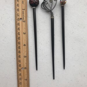 May include: Three black hair sticks with decorative tops. One has a red and black speckled ball, another has a silver filigree design with rhinestones, and the third has a gold and crystal ball. A wooden ruler is next to the hair sticks.