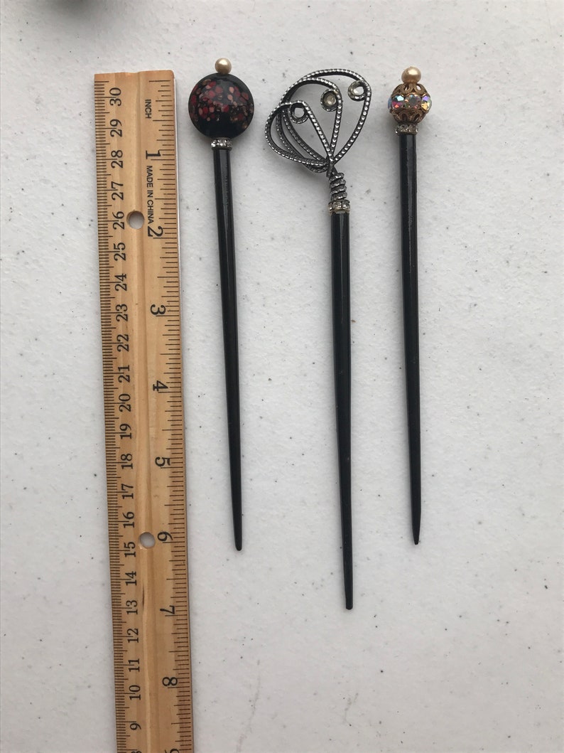 May include: Three black hair sticks with decorative tops. One has a red and black speckled ball, another has a silver filigree design with rhinestones, and the third has a gold and crystal ball. A wooden ruler is next to the hair sticks.