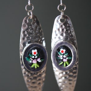 May include: Silver earrings with a hammered finish and a black oval pendant featuring a mosaic floral design with white, pink, and green accents.