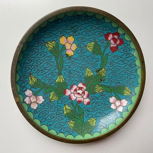May include: A small, round, blue and green enamelware dish with a floral design. The dish features pink, yellow, and red flowers with green leaves and stems. The background is a light blue with a pattern of white clouds.