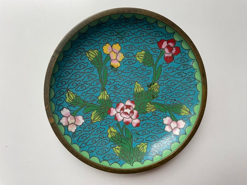 May include: A small, round, blue and green enamelware dish with a floral design. The dish features pink, yellow, and red flowers with green leaves and stems. The background is a light blue with a pattern of white clouds.