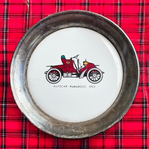May include: A round metal tray with a white center featuring a red vintage car illustration. The car is labeled "AUTOCAR RUNABOUT, 1902". The tray has a silver-colored rim and sits on a red and black plaid surface.