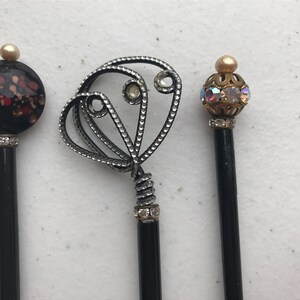 May include: Three decorative pencils with unique toppers. One has a black and red floral design, another features a silver filigree heart, and the third has an iridescent orb. All have black shafts and rhinestone accents.