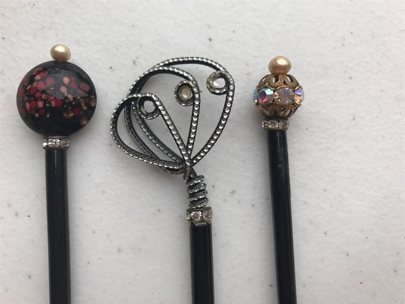 May include: Three decorative pencils with unique toppers. One has a black and red floral design, another features a silver filigree heart, and the third has an iridescent orb. All have black shafts and rhinestone accents.