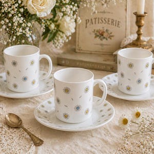 May include: Three white ceramic teacups and saucers with a daisy pattern. The cups have a handle and are placed on matching saucers. A vintage gold spoon rests on the table. The background includes flowers and a decorative sign.