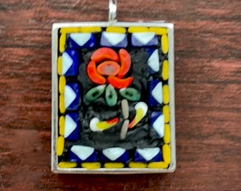 May include: A square mosaic pendant with a silver frame. The mosaic features a red flower with green leaves on a black background. The frame has a yellow and blue border.