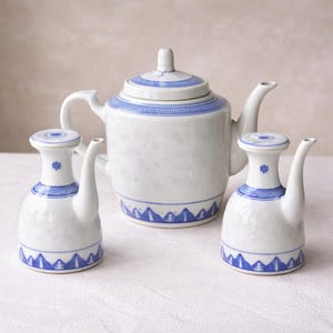 May include: A white porcelain tea set with blue decorative trim. The set includes a teapot with a lid and two small pitchers. The blue trim features geometric and floral patterns. The tea set is displayed on a white surface.