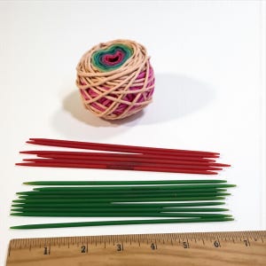 May include: A still-life image of knitting supplies. A ball of yarn with pink, green, and tan hues sits above two sets of knitting needles. The top set is red, and the bottom set is green. A wooden ruler is at the bottom.