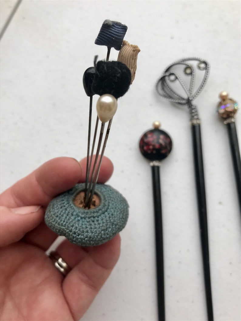 May include: A collection of vintage hat pins. One pin is held in a hand, featuring a blue crocheted base with various decorative tops, including a pearl, black velvet, and fabric accents. Other pins with black shafts and decorative tops are also visible.