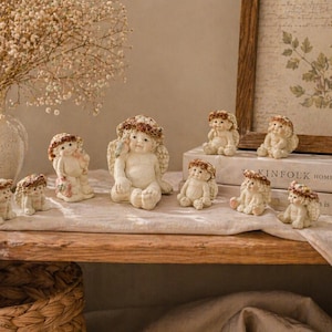 May include: A collection of small, cream-colored angel figurines with detailed wings and floral headbands. The angels are arranged on a wooden shelf with a neutral-toned background, alongside a vase of dried flowers and a framed print.