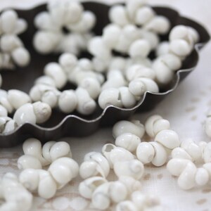 May include: A collection of small, white, spiral-shaped seashells, resembling tiny conch shells, are scattered around a dark, scalloped-edge metal dish. The shells are clustered together, creating a textured, organic pattern. The background is a soft, off-white fabric.
