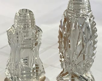 May include: Two clear glass salt and pepper shakers with intricate cut glass designs. The shakers are both tall and slender, with a hexagonal base.