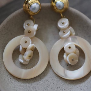 Könnte beinhalten: Ein Paar goldene und weiße Muschelohrringe mit Perlenakzent. Die Ohrringe haben einen großen Muschelkreis mit einem kleineren Cluster von Muschelkreisen, die daran hängen.