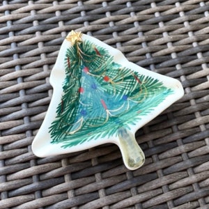May include: A white ceramic Christmas tree-shaped dish with a painted green tree design. The tree is decorated with red and blue ornaments, gold garland, and a gold star on top. The dish has a small handle.