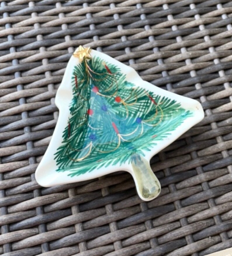 May include: A white ceramic Christmas tree-shaped dish with a painted green tree design. The tree is decorated with red and blue ornaments, gold garland, and a gold star on top. The dish has a small handle.