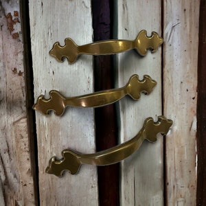 May include: Three antique brass drawer pulls with ornate designs. The pulls are mounted on a weathered wooden surface.