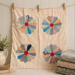 May include: A cream-colored textile with four large, colorful flower designs. Each flower has a red center and radiating petals in various patterned fabrics. The textile is hung with wooden clothespins against a white wall.