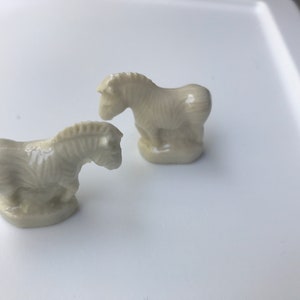May include: Two small white ceramic zebra figurines. The zebras are standing on a white surface.