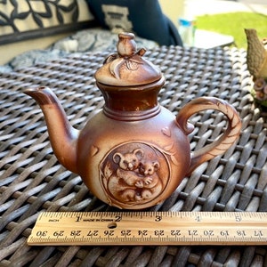 May include: A small brown ceramic teapot with a koala bear design. The teapot has a lid with a flower on top. The teapot is 3 inches tall and is made in China.