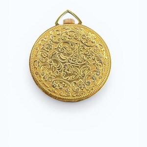 May include: A gold-colored, round pocket watch with an ornate floral design. The watch has a loop at the top for a chain. The intricate details of the design are visible across the entire surface of the watch.
