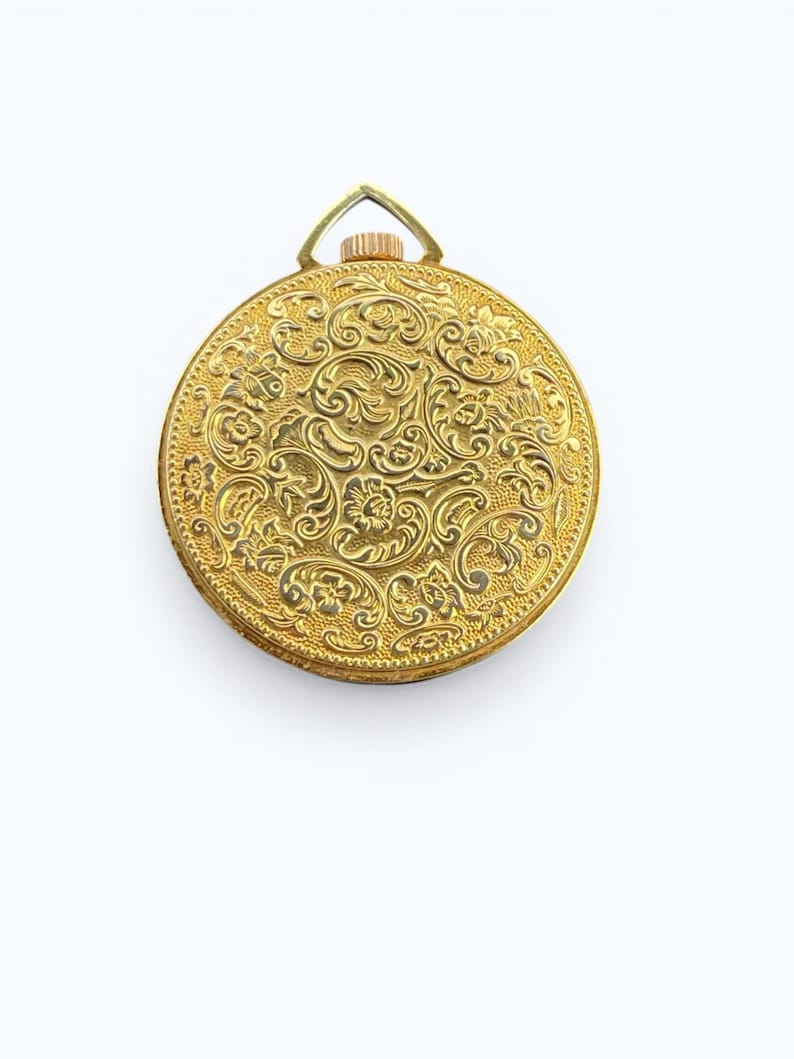 May include: A gold-colored, round pocket watch with an ornate floral design. The watch has a loop at the top for a chain. The intricate details of the design are visible across the entire surface of the watch.