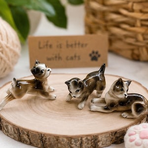 May include: Three ceramic cat figurines in brown and white, posed on a wooden slice. One cat is standing, two are in a playful position. A small card reads "Life is better with cats" with a paw print.