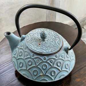 May include: A teal-colored cast iron teapot with a black handle. The teapot features a textured, patterned design on the body and lid. The handle is arched, and the spout is short and curved. The teapot is sitting on a dark wooden surface.