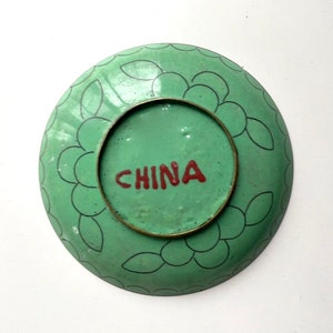 May include: A green and black enamelware plate with a floral design. The plate has a brass rim and the word "CHINA" written in red on the bottom.