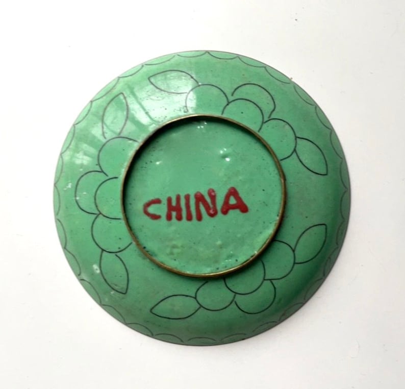 May include: A green and black enamelware plate with a floral design. The plate has a brass rim and the word "CHINA" written in red on the bottom.