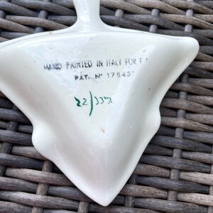 May include: A white, triangular ceramic dish with a handle, hand-painted in Italy. The dish has a unique shape and features text that reads "HAND PAINTED IN ITALY FOR F..." and "PAT. N&deg; 17843". The number "22/335" is also visible.