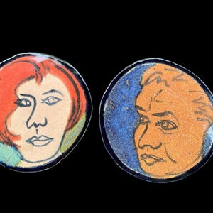 May include: Two round ceramic pieces with painted faces. One features a woman with red hair and a light skin tone, the other a man with orange skin and a blue background. Both have black outlines.