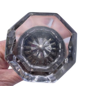 May include: Clear, octagonal glass doorknob with a faceted design. The knob has a central, circular pattern and is held against a white background. The glass reflects light, creating highlights and shadows.