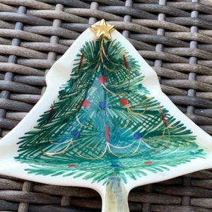 May include: A Christmas tree-shaped decorative dish. The ceramic dish features a hand-painted Christmas tree design with green, blue, and red accents, topped with a gold star. The dish is white with a textured surface.