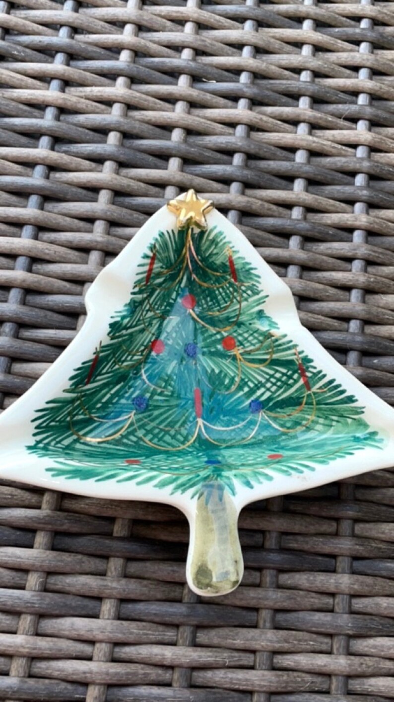 May include: A Christmas tree-shaped decorative dish. The ceramic dish features a hand-painted Christmas tree design with green, blue, and red accents, topped with a gold star. The dish is white with a textured surface.