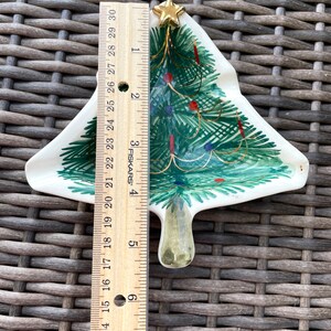May include: A ceramic Christmas tree-shaped dish, approximately 6 inches tall, with green painted branches, red and blue ornaments, and a gold star on top. The dish has a white background and is on a woven surface.