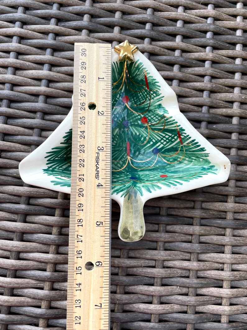 May include: A ceramic Christmas tree-shaped dish, approximately 6 inches tall, with green painted branches, red and blue ornaments, and a gold star on top. The dish has a white background and is on a woven surface.