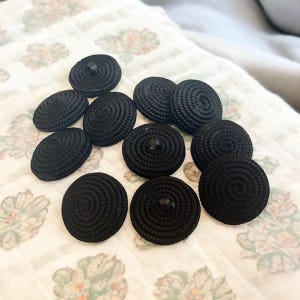 Pu&ograve; includere: Una collezione di 10 bottoni rotondi neri con un design a spirale. I bottoni sono realizzati in tessuto intrecciato e hanno un retro piatto.