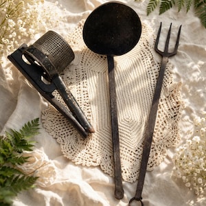 May include: An assortment of antique kitchen tools, including a potato ricer, a large round ladle, and a long-handled fork. The tools are made of dark metal and rest on a cream-colored crocheted doily and linen cloth, with sprigs of white flowers and ferns.