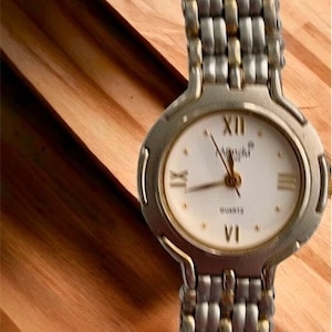 May include: A silver and gold watch with a white face and black Roman numeral hour markers. The watch has a quartz movement and is on a silver and gold bracelet.