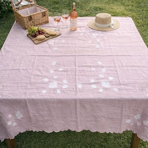 May include: A picnic scene with a pink tablecloth featuring white floral embroidery. A wicker basket, wine glasses, a bottle of rosé, and a straw hat are arranged on the table. A wooden cutting board with grapes and crackers is also present.