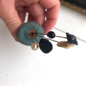 May include: A collection of antique hat pins with various decorative heads. The pins are attached to a blue pin cushion. The pin heads include a black bead, a pearl, a navy blue fabric design, and a tan fabric design.