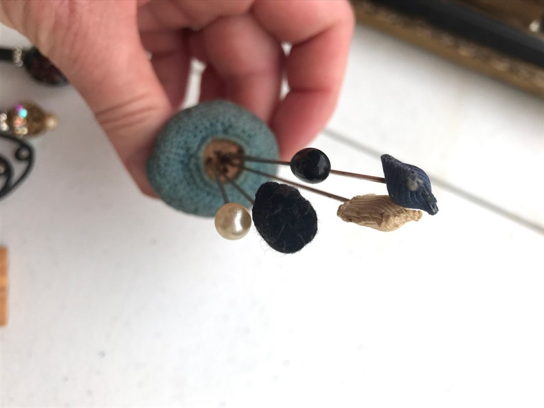 May include: A collection of antique hat pins with various decorative heads. The pins are attached to a blue pin cushion. The pin heads include a black bead, a pearl, a navy blue fabric design, and a tan fabric design.