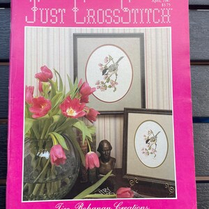 May include: A pink cover of "Just CrossStitch" magazine with a floral arrangement and framed cross-stitch art. The magazine cover features the title in a stitched font, with the date March/April 1987. The artwork depicts a bird on a flowering branch.