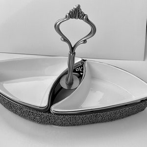 May include: A white ceramic serving dish with two sections and a decorative metal handle. The dish has a detailed, intricate design around the rim.