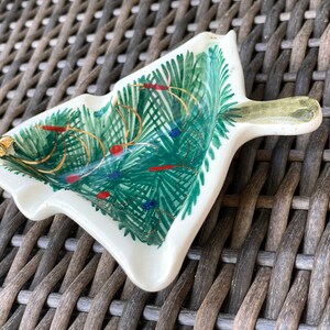May include: A ceramic Christmas tree-shaped dish with a handle. The dish is painted with green fir needles, gold garland, and red and blue ornaments. A gold star adorns the tip of the tree.