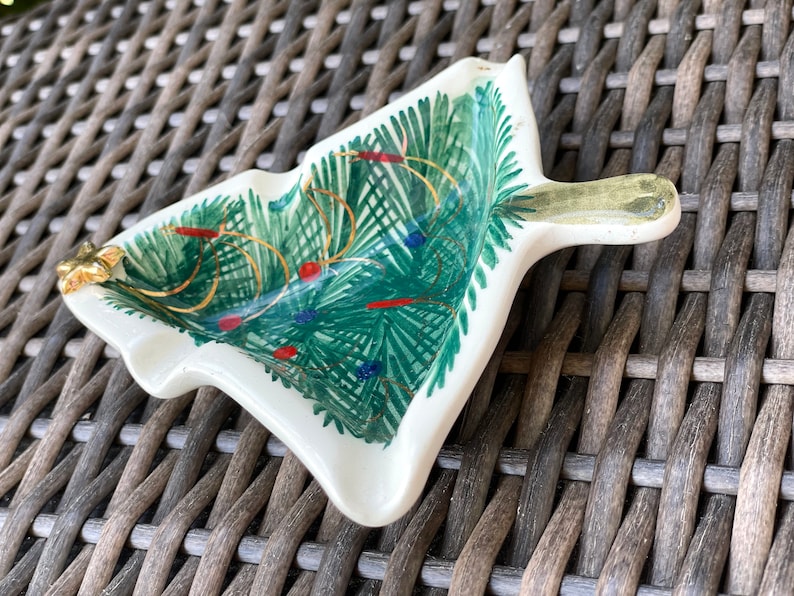 May include: A ceramic Christmas tree-shaped dish with a handle. The dish is painted with green fir needles, gold garland, and red and blue ornaments. A gold star adorns the tip of the tree.