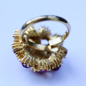 May include: A gold ring with a large, red gemstone surrounded by smaller red gemstones. The ring is set in a gold band.