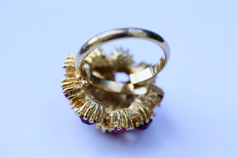 May include: A gold ring with a large, red gemstone surrounded by smaller red gemstones. The ring is set in a gold band.